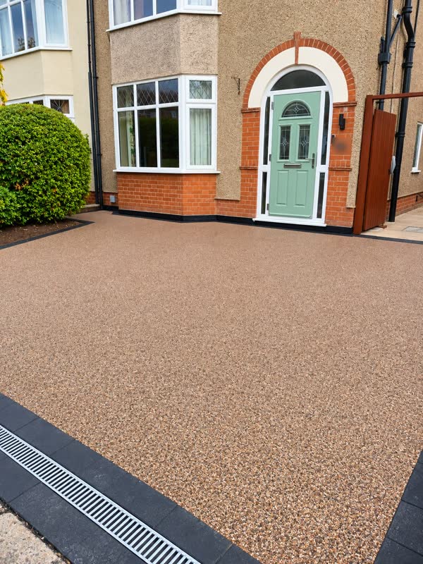 Warm-toned resin bound driveway in front of a bay-windowed house, with black block edging and a drainage channel